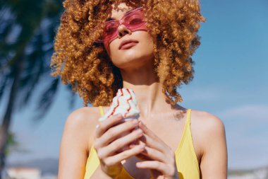 Neşeli bir kadın dondurma tutuyor, pembe güneş gözlüğü ve sarı bir bluz takıyor, neşe ve yaz havasını yansıtıyor.