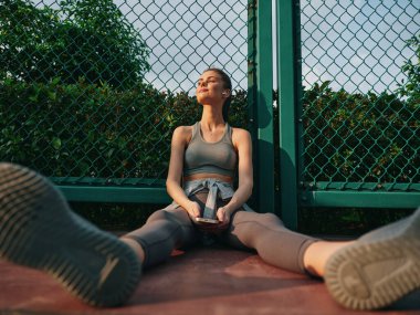 Genç bir bayan spor meraklısı, elinde bir telefonla mahkeme salonunda oturuyor. Etrafı yeşil yapraklarla çevrili. Sükunet ortamında.