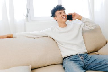 Modern ve parlak bir oturma odasında, yumuşak bir kanepede uzanan mutlu genç adam doğal ışıkla dolu, bir anlık neşe ve bağlantı yakalıyor.