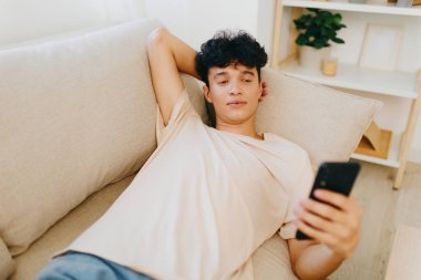 Genç adam bej bir kanepeye uzanmış, akıllı telefonuna odaklanmış, rahat bir ev atmosferi sergiliyor arka planda nötr renkler ve çağdaş dekor unsurları var, yaşam tarzı tasviri için ideal.