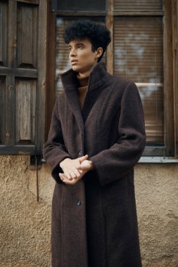 Kahverengi ceketli moda adamı pencerenin önünde duruyor, elleri kalçasında, kameradan uzağa bakıyor..