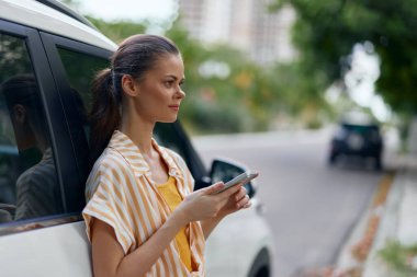 Genç bir kadın park halindeki bir arabanın yanında elinde akıllı bir telefonla dikiliyor ve düşünceli bir şekilde ekrana bakıyor. Üzerinde çizgili bir kıyafet var, yeşil bir şehir geçmişine sahip.