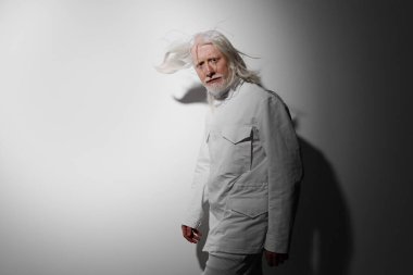 Beyaz ceketli, uzun beyaz saçlı yaşlı bir adamın portresi yumuşak gri bir arka plana yaslanmış, dingin ve sanatsal bir varlık yayıyor.