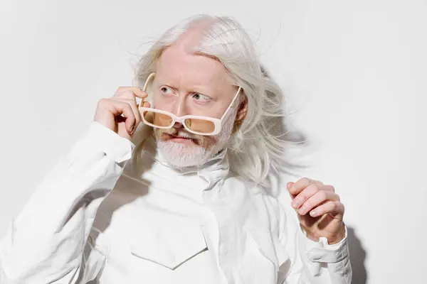 stock image A striking portrait featuring an elderly man with long white hair and beard, he is adjusting his unique glasses while wearing a contemporary white outfit The clean, bright background highlights his