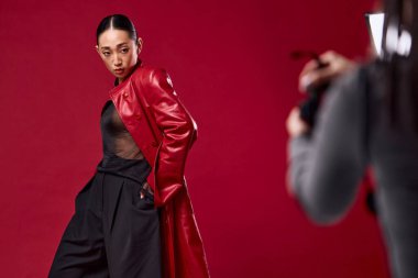 Kırmızı deri ceketli ve pantolonlu bir kadınla Asyalı bir moda çekimi kırmızı arka planda poz verirken bir fotoğrafçı onun resmini çekiyor..