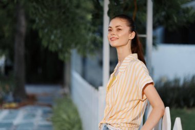 Sarı ve beyaz çizgili bir gömlek giymiş, neşeli bir ifadeyle genç bir kadın, beyaz bir çitin yanında duruyor, yemyeşil bir çitle çevrili, doğal bir huzur ve memnuniyet hissi yayıyor.