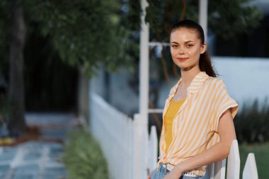 Genç bir kadın, çizgili şık, rahat bir kıyafetle, yeşil bir zemin üzerinde duruyor. Sıcak doğal ışık, onun özelliklerini vurguluyor, bir huzur ve mutluluk hissi veriyor.