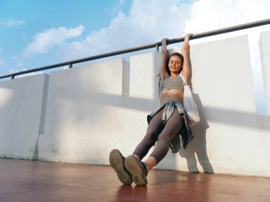 Formda bir bayan atlet çatıda asılma egzersizi yapıyor. Parlak ve motive edici bir ortamda güç ve kararlılık gösteriyor.