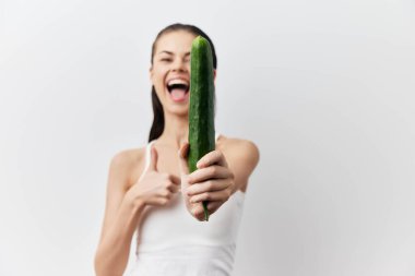 Hafif kıyafetli neşeli genç bir kadın, salatalığı tutarken gülümseyen ve baş parmağını kaldıran, düz beyaz bir arka plan üzerine kurulmuş.