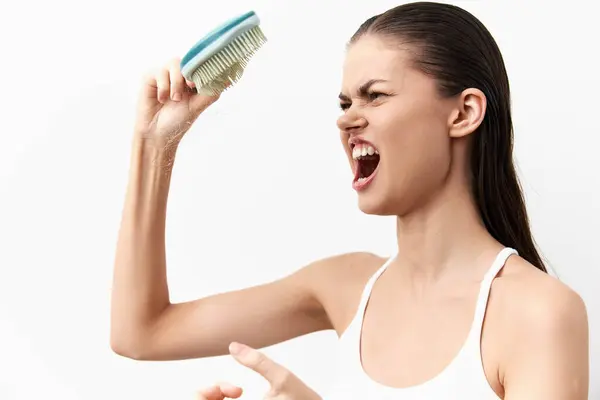 stock image A young woman showcasing intense emotion and frustration with an aggressive expression while gripping a brush She is dressed in a plain white tank top, set against a bright white background