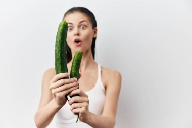 Şaşırtıcı bir ifadeyle iki yeni seçilmiş salatalığı minimalist beyaz arka planda tutan şaşkın bir kadın mizahi ve neşeli bir an yaratıyor.