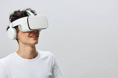 Genç adam sanal gerçeklik deneyiminden zevk alıyor, parlak bir VR kulaklık ve kulaklık takarken gülümsüyor. Minimalist açık gri arka plan gelişmiş teknoloji ve dalkavukluğu vurguluyor.