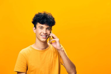 Kıvırcık saçlı neşeli genç bir adam, turuncu bir tişört giymiş, çarpıcı sarı bir arkaplan üzerine kurulmuş bir telefon görüşmesi yapıyor.