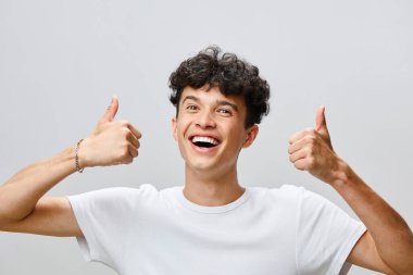 Kıvırcık saçlı, beyaz tişörtlü mutlu genç adam, coşkuyla baş parmak kaldırma hareketi sergiliyor yumuşak gri arka plan onun neşeli ifadesini vurguluyor ve temasını güçlendiriyor