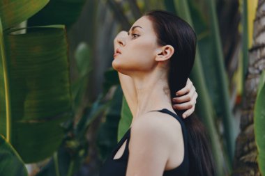 Uzun siyah saçlı kadın, güneşli bir günde ağaçların önünde palmiye ağaçlarıyla dikiliyor.