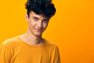Koyu kıvırcık saçlı mutlu bir genç adam, canlı turuncu bir arkaplan karşısında kameraya güvenle bakıyor. Günlük sarı tişörtü görüntüyü canlandırıyor.