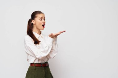 surprised young woman dressed in a white shirt and green trousers, standing against a light grey background, displays excitement with an open mouth and hand gesture, conveying emotions vividly clipart