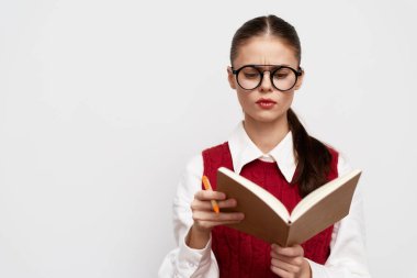 Gözlüklü genç bir kadın elinde turuncu bir kalemle kahverengi bir kitap okumaya odaklanmış. Kırmızı yelek ve beyaz bir gömlek giymiş. Düz bir arkaplanda duruyor.