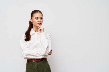 Beyaz gömlek ve yeşil pantolon giyen genç bir kadın eli çenesinde, beyaz bir arkaplan önünde düşünceli bir şekilde duruyor. Düşünce ve içgözlem hissini aktarıyor.