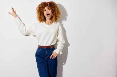 Şehvetli kıvırcık saçlı, rahat beyaz bir kazak ve yüksek bel kot giyen, açık renkli arka planda duran neşeli bir kadın, coşku ve tazeliği iletiyor.