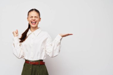 Neşe ve memnuniyeti ifade eden mutlu bir kadın şık bir beyaz gömlek ve yeşil pantolon giyer, kişisel başarısını kutlar, tarafsız açık gri bir arka plana karşı, Po 'yu taşımak için ideal