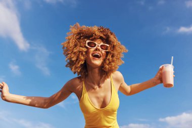 Güneş gözlüğü takan neşeli genç bir kadın ve sarı bir mayo elinde ferahlatıcı bir içecek, açık havada mutluluk yayıyor.