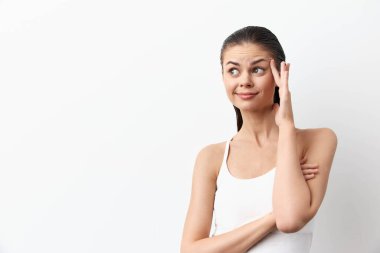 Beyaz atletli genç bir kadın temiz beyaz bir zemine karşı duruyor, düşünceli bir tavır sergiliyor. Gülümserken alnına dokunuyor, merakını iletiyor ya da kafa yoruyor.