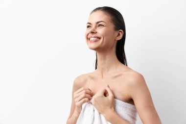 Islak saçlı neşeli bir genç kadın, bir havluya sarılmış, ışıl ışıl gülümsüyor, temiz beyaz bir arkaplan önünde duruyor, tazelik ve kendine güven yayıyor.