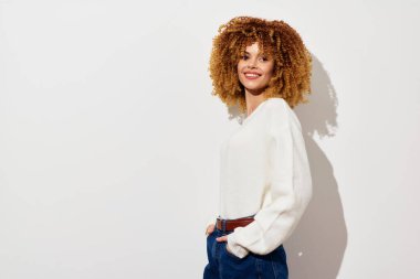 Kıvırcık saçlı neşeli bir kadın, rahat bir poz veren beyaz bir süveter giyer. Yumuşak beyaz arka plan, neşeli ifadesini ve şık kıyafetini vurgular.