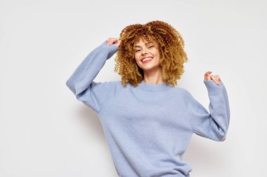 Kıvırcık saçlı, yumuşak mavi bir kazak giyen, beyaz bir arkaplanda dans eden ve gülümseyen neşeli genç kadın görüntüsü mutluluk, özgürlük ve kendini ifade ediyor.