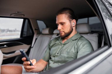 Yeşil gömlekli bir adam arabanın arka koltuğunda oturuyor, telefonuyla meşgul, modern teknoloji ve yaşam tarzını gösteriyor.