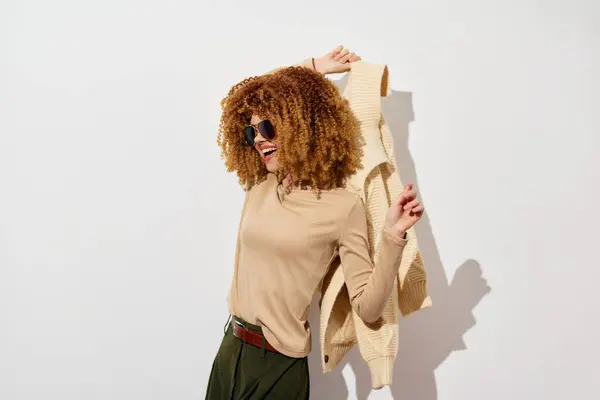 stock image Stylish young woman with curly hair is playfully posing against a clean white background She wears a beige top and sunglasses, exuding a cheerful and confident vibe