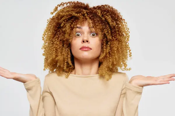 stock image surprised woman with curly hair expressing confusion, looking directly at the camera with an open mouth She raises her hands in a gesture of uncertainty against a soft, light background