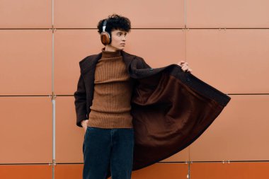 Kahverengi ceketli ve kulaklıklı bir adamın, elleri kalçaları ve kolları açık, turuncu bir duvara poz verirken moda fotoğrafı..