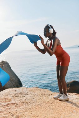 Kırmızı kıyafetli fit bir kadın, sakin bir su ve açık bir gökyüzü ile çevrili kayalık bir sahilde mavi yoga minderiyle pratik yapıyor.