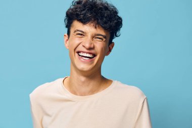Kıvırcık saçlı neşeli bir genç, parlak mavi bir arka planda geniş bir gülümseme sergiliyor. Açık renk gömleği parlak atmosferi tamamlıyor. Neşeli tavırlarını ve sıcak enerjisini vurguluyor.