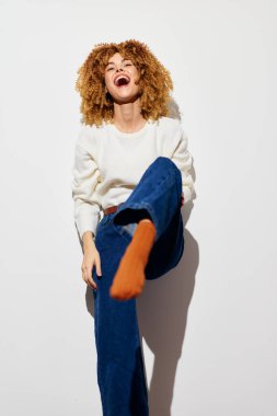 Kıvırcık saçlı neşeli bir kadın, neşeli bir şekilde bacağını kaldırırken gülüyor. Görüntü, temiz beyaz bir arka planda kaygısız bir ruh yakalıyor. Mutluluğu vurguluyor.