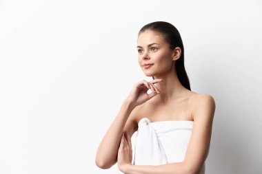 Beyaz bir havluya sarılı genç bir kadın, sade beyaz bir zemin üzerinde düşünceli bir şekilde poz veriyor, sakinlik ve kendine güven duygusu somutlaştırıyor, güzellik ve sağIık teması için mükemmel.