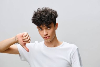 Beyaz tişörtlü, kıvırcık saçlı, hayal kırıklığına uğramış genç bir adam yumuşak gri bir arka plana başparmak işareti yapıyor. Yüzündeki ifade hayal kırıklığını yansıtıyor ve uygun bir atmosfer yaratıyor.