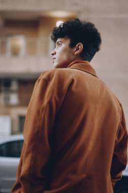 Kıvırcık saçlı, şık kahverengi ceketli, şehir ortamında dikkatle bakan genç bir adam.