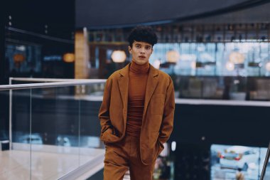 Kahverengi takım elbise ve balıkçı yaka giyen zarif ışıklandırma ve mimarisi ile modern bir iç mekanda yürüyen kendine güveni ve entelektüelliği olan şık bir genç.