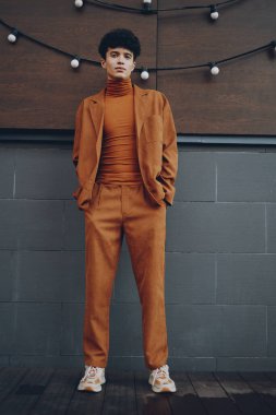 Turuncu takım elbiseli ve balıkçı yakalı şık bir erkek model, yumuşak ışıklandırmalı, kendinden emin ve modern stili olan şık ahşap bir duvara karşı duruyor.