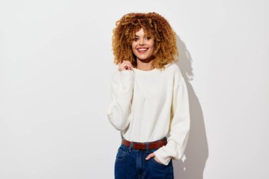 Bu resim, şehvetli kıvırcık saçlı, sıradan beyaz bir süveter giymiş neşeli bir genç kadını gösteriyor. Basit beyaz bir arkaplanda duruyor, pozitifliği ve yaklaşılabilirliği yansıtıyor, mükemmel.