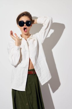 Beyaz gömlek ve zeytin yeşili etek giyen şık manken, modern trendleri ve hayat dolu yaşam tarzını temsil eden temiz beyaz bir arka plana karşı büyük ölçekli güneş gözlükleriyle şaşkınlığını ifade ediyor.