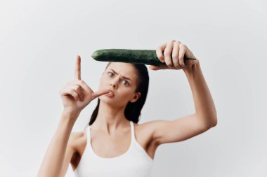 Uzun siyah saçlı genç bir kadın bir elinde bir salatalık tutarken diğer eliyle bir L şekli oluşturuyor. Mizahi ifadesi, temiz beyaz bir zemin üzerine kurulmuş neşeli bir tavır sergiliyor.