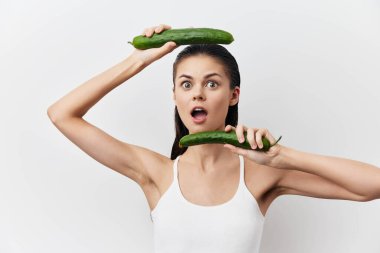 Genç bir kadın dramatik bir şekilde salatalıkları başının üstünde ve yüzünün önünde temiz beyaz bir zemin üzerinde tutar, şaşkın ifadesini ve neşeli ve parlak kişiliğini vurgular.