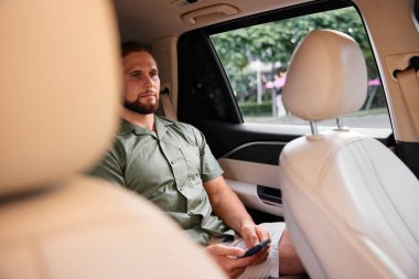 Bir arabanın içinde oturmuş, akıllı telefonuna odaklanmış, rahat giyinmiş genç bir adam açık renk, pencereden görünen yeşil yapraklarla çelişiyor.