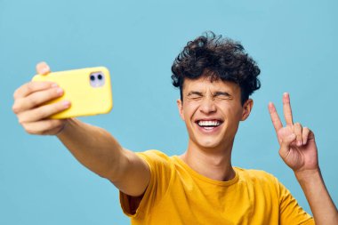 Kıvırcık saçlı neşeli genç bir adam canlı mavi bir arkaplan karşısında akıllı telefonuyla selfie çekiyor. Parlak sarı bir tişört giyiyor ve barış işareti yapıyor.