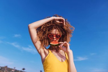 Canlı bir yaz sahnesi. Sarı mayo giymiş bir kadın. Bir dilim karpuzla neşeli bir poz veriyor.