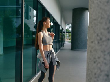 A fit woman in active wear jogs outdoors, smiling with headphones on, in an urban setting featuring large glass windows